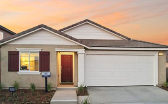 A modern, single-story house with a tan exterior and dark red shutters. It has a double garage, a well-lit entrance, and a sunset sky background.