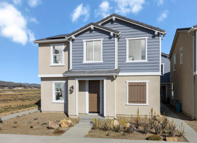 A two-story house with beige walls and blue siding, featuring large windows, a brown front door, and a landscaped yard with sparse vegetation and rocky terrain.