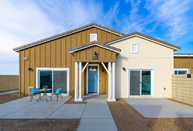 Modern residential home with a brown and white facade, featuring a blue door, large windows, and a patio with turquoise chairs.